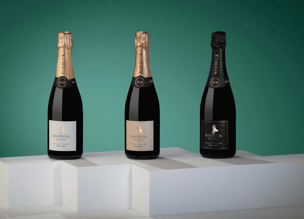 Three bottles of Roebuck Estates English sparkling wine, Classic Cuvée 2018, Rosé de Noirs 2019, and Blanc de Noirs 2018, are elegantly arranged on white plinths against a green background